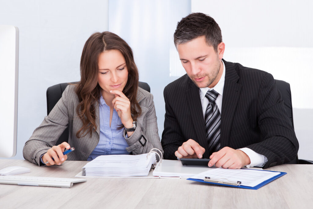 Two Young Businesspeople Calculating Bills At Desk In Office