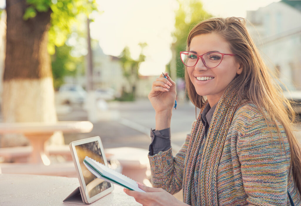 Pretty stylish hipster woman in glasses sitting on street with tablet and notepad having studies and smiling at camera